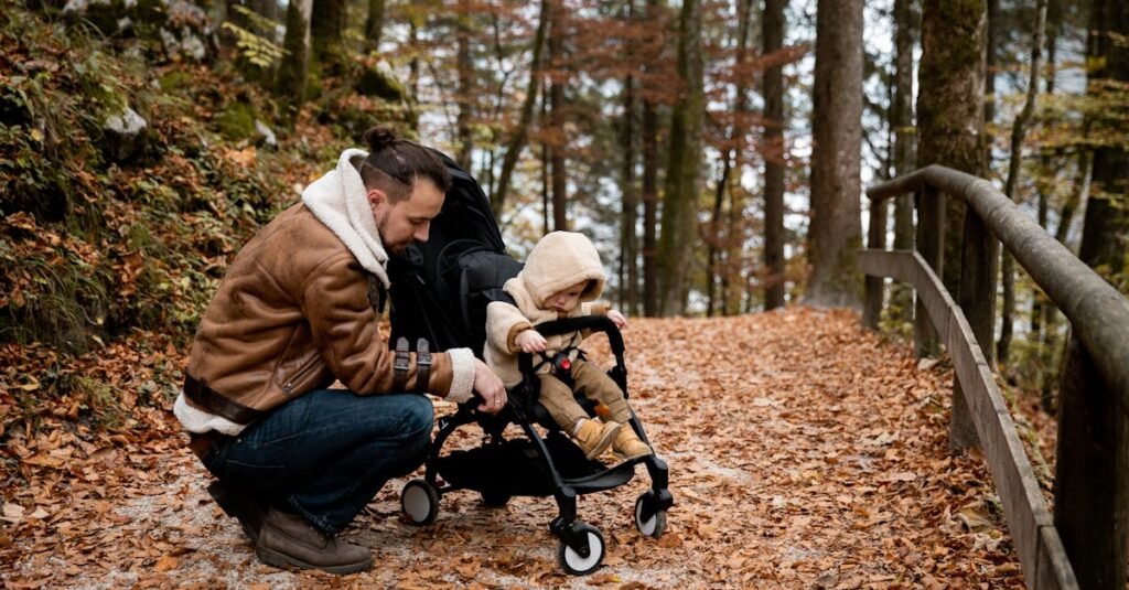 A father kneels beside his toddler in a stroller, amidst a scenic autumn forest path.